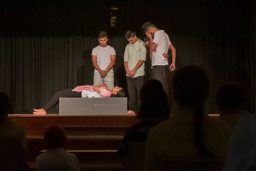 View from behind the heads of people of a stage with young people acting sad and a girl lying down, acting dead or very ill
