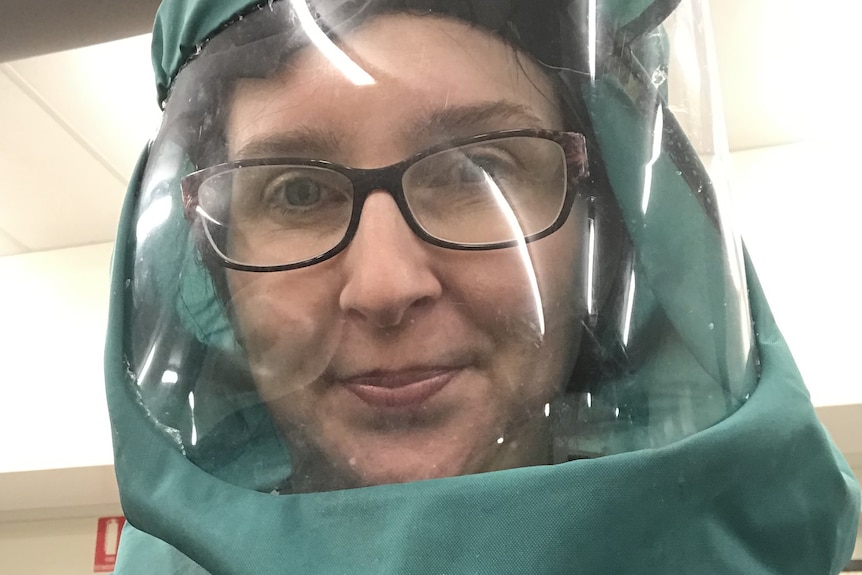 Woman in green hazmat suit with full clear face mask and respirator, she is wearing glasses and smiling