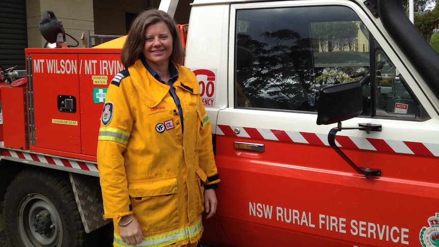 A smiling woman with dark, shoulder length hair, dressed in a firefighting uniform and standing in front of a fire truck.