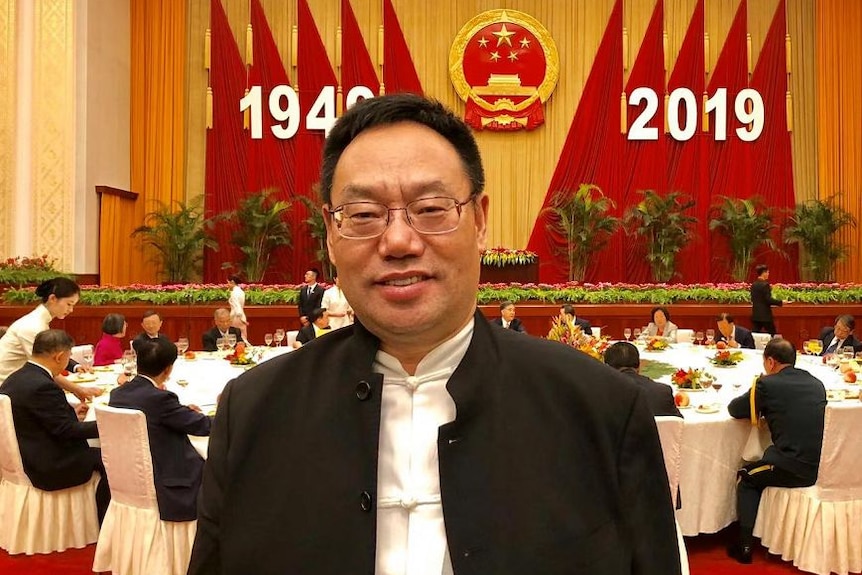 A man in formal attire poses in front of a banner featuring a Chinese government crest and the years 1949 and 2019.