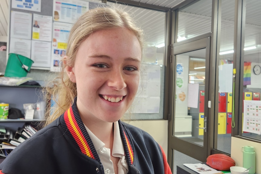 A blond haired teenager in school uniform smiles at the camera