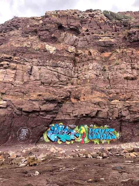 A large patch of colourful graffiti on a rock wall at the bottom of a cliff