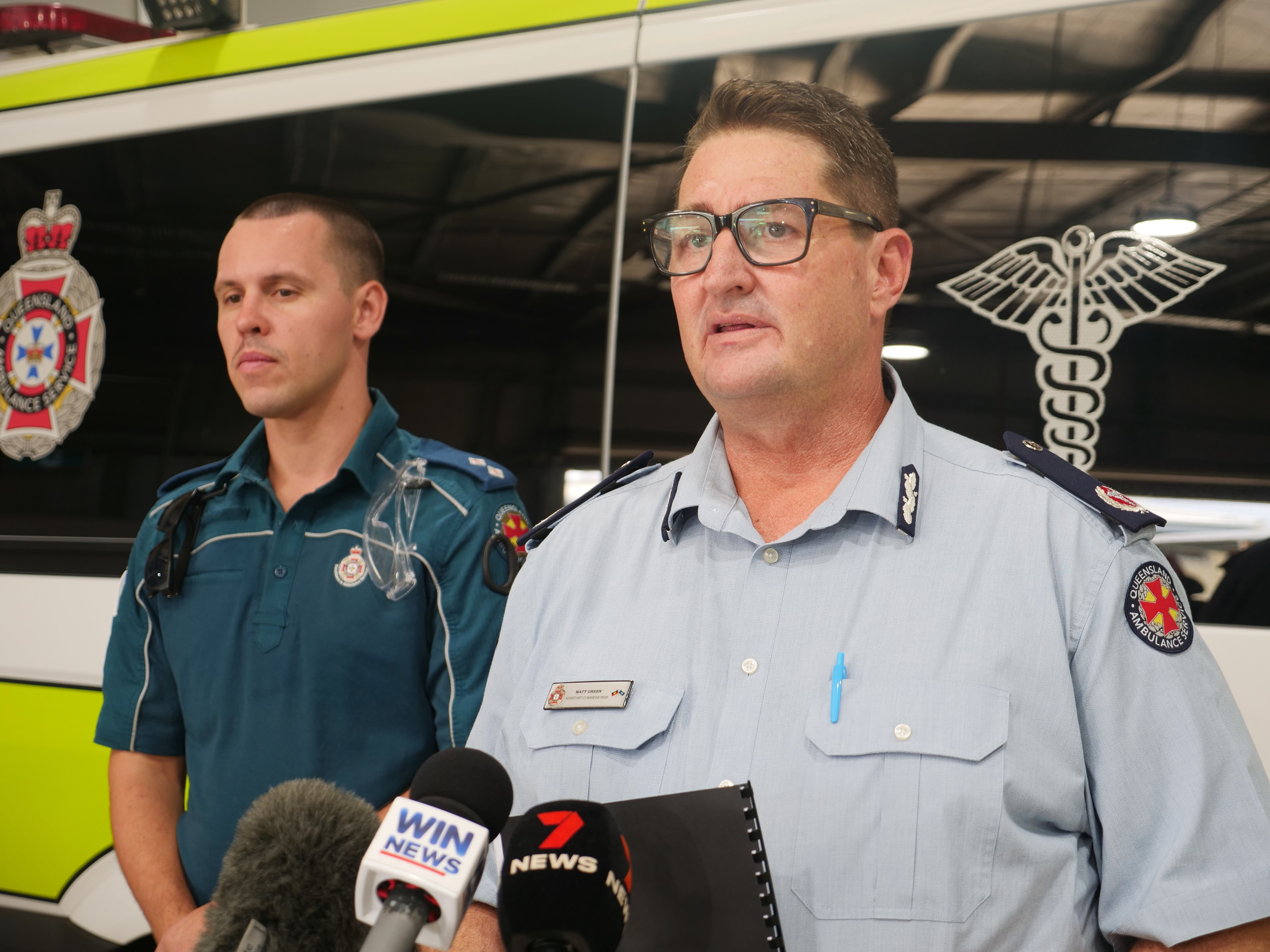 Two paramedics in uniform stand before an ambulance vehicle. They both look serious. 