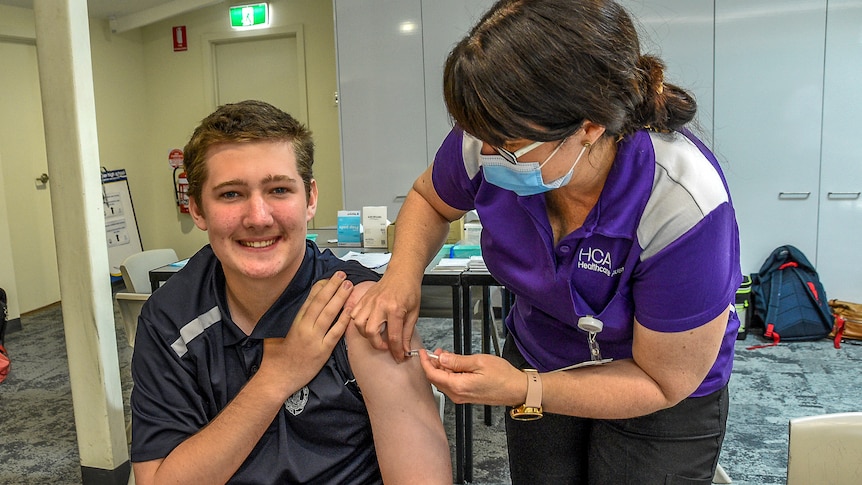 A boy in a school uniform smiles as a nurse slides a vaccine needle into his arm.