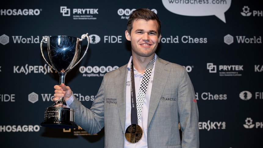 A smiling man holds a trophy in the air while he has a medal around his neck after successfully defending the world chess title.