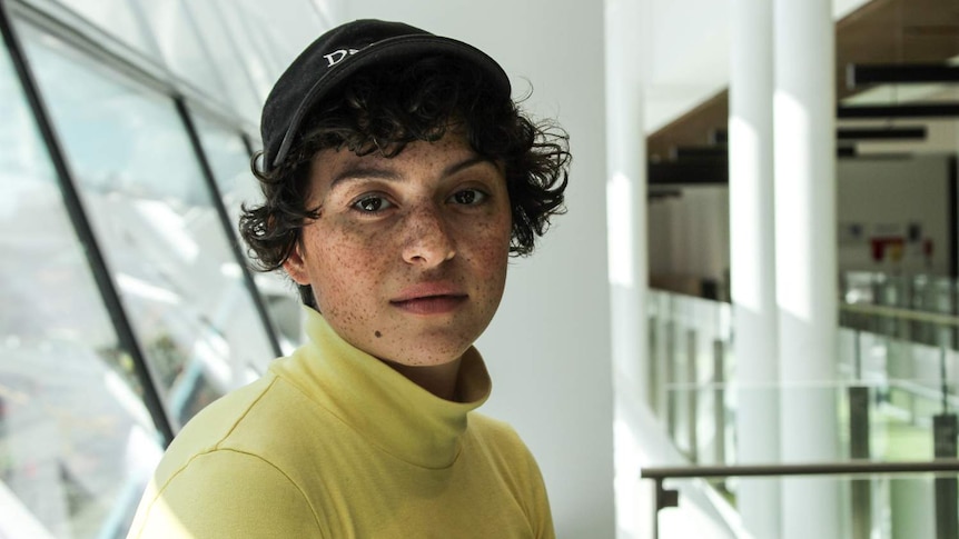 Actress with short curly haircut wearing black baseball cap and lemon-yellow turtle neck top, looking at camera