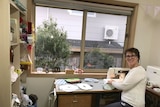 Tracey Nuthall sits at her desk by a window with a sewing machine, holding material and smiling at the camera.
