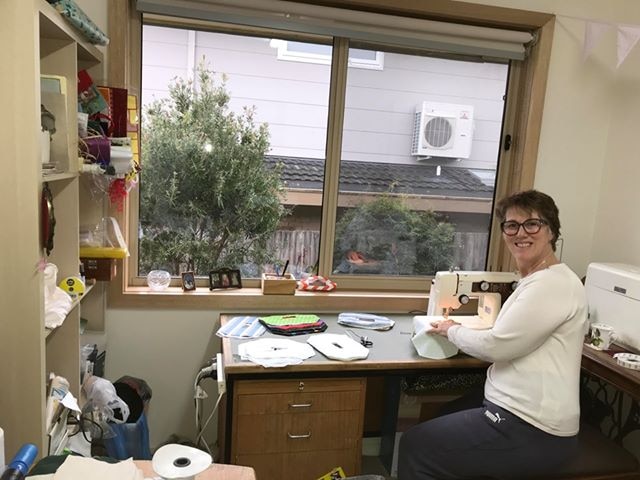 Tracey Nuthall sits at her desk by a window with a sewing machine, holding material and smiling at the camera.