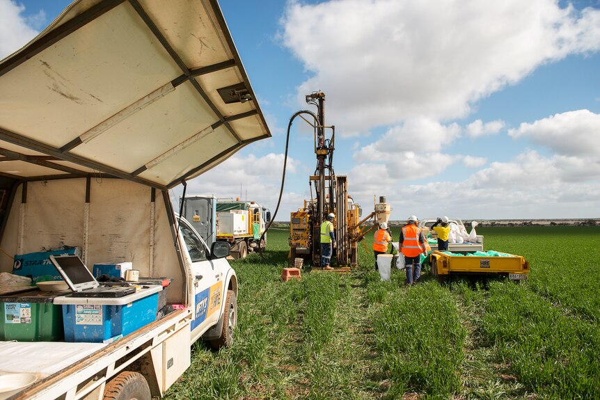 Four mine workers in high-vis uniforms stand between a ute and mining machinery in a cereal paddock.