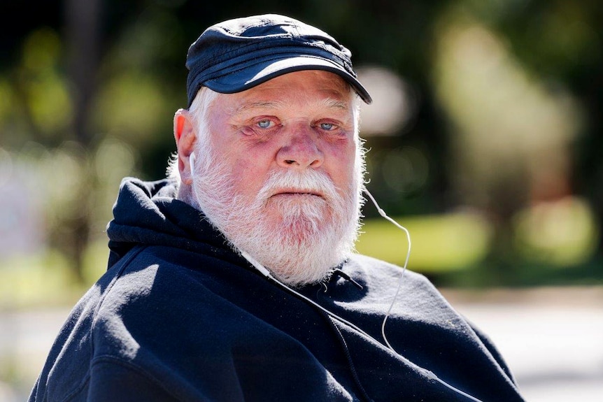 A head and shoulder shot of an older man with a white beard wearing a black cap and jumper and headphones.