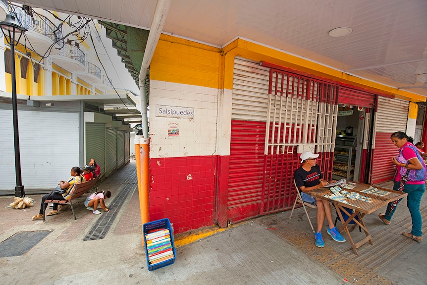 Older man sits behind a card table on a street corner selling lotto tickets.