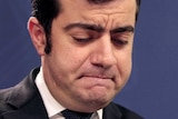 Sam Dastyari looks down, frowning, standing at a microphone. There is an Australian flag behind him.