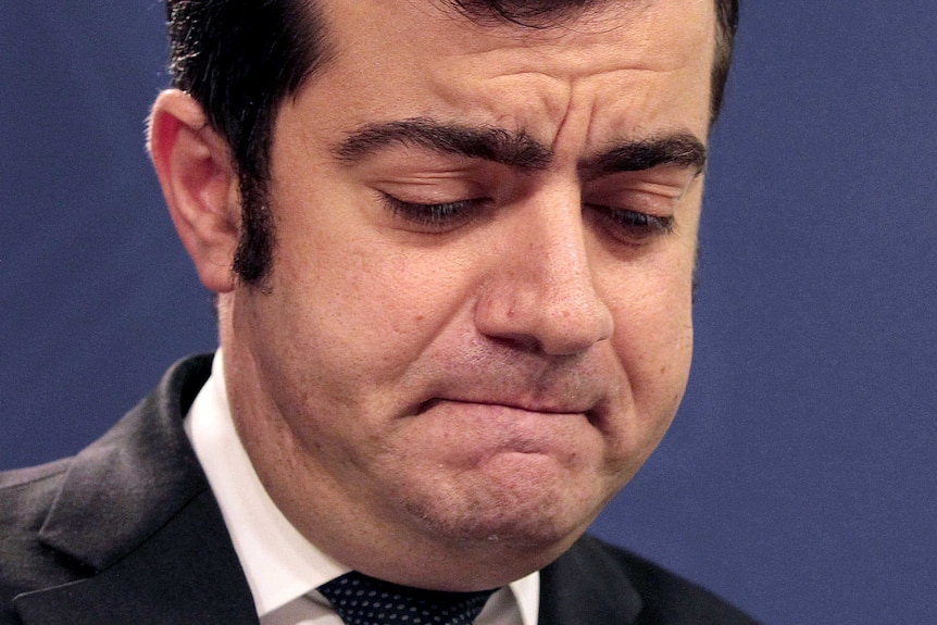 Sam Dastyari looks down, frowning, standing at a microphone. There is an Australian flag behind him.