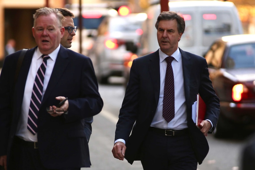 Two men walking down a city street wearing suits, with a third man in the rear.