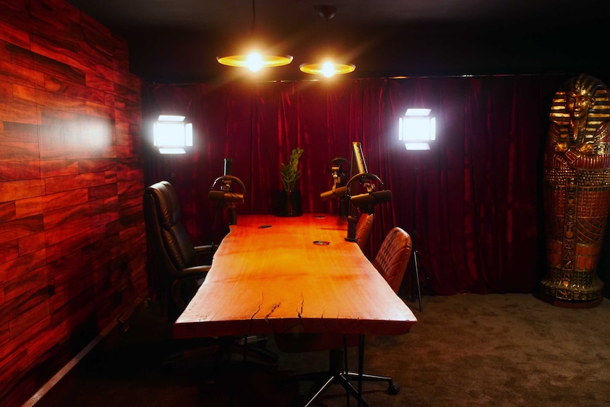 A long wooden table in the middle of a room with recording studio equipment, a red velvet wall and an Egyptian mummy statue.
