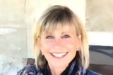 Still from video showing Olivia Newton-John looking healthy and smiling widely, sitting outside on a chair.