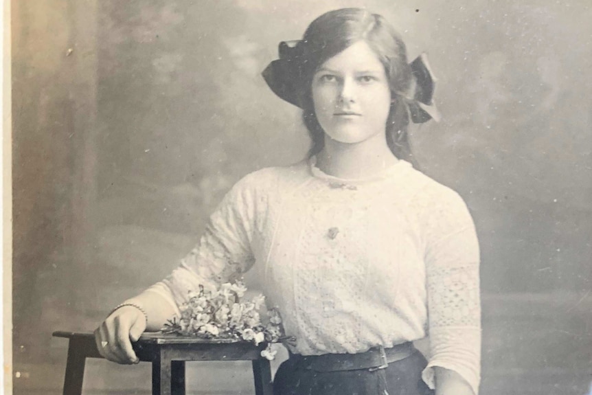 A black and white photograph of a woman leaning on a timber stool with flowers on it.