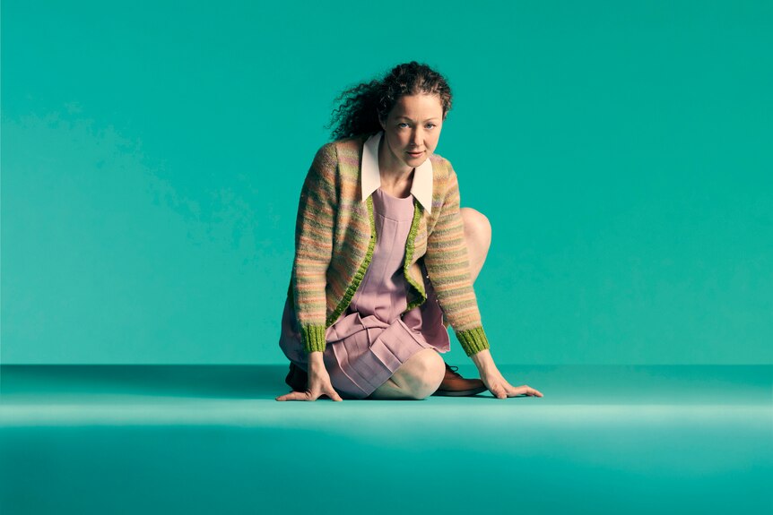 A curly-haired woman in her 30s wearing a cardigan and 60s-style dress is crouched on a turquoise floor
