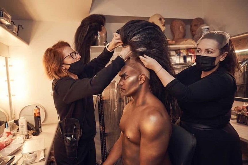 A Torres Strait Island man, shirtless with heavy make-up, has a large mane-like wig attached by two women backstage at a theatre