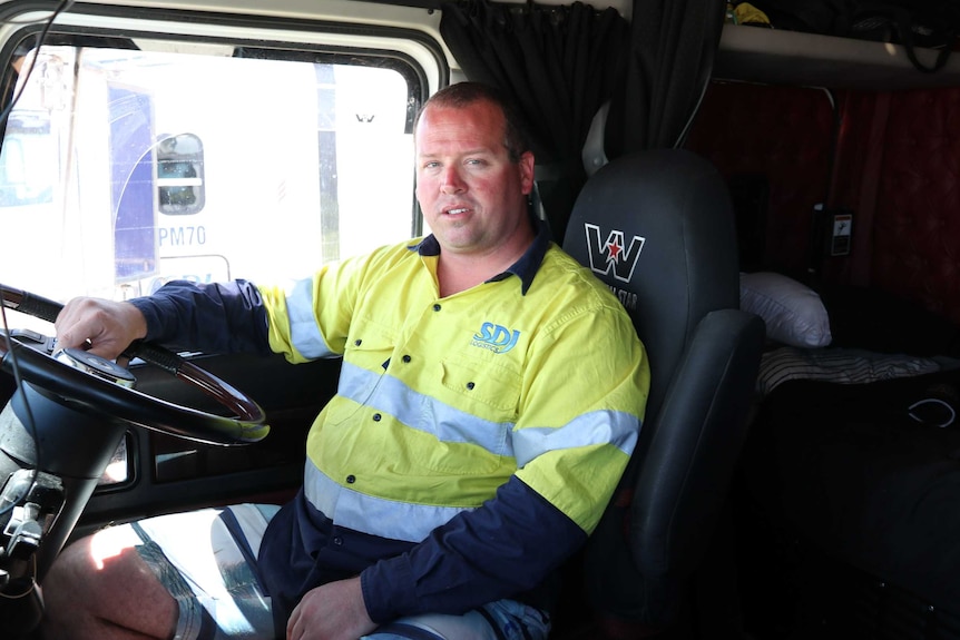 A middle aged man wearing a high visibility work shirt sits in the cabin of a large truck.