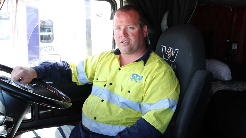 A middle aged man wearing a high visibility work shirt sits in the cabin of a large truck.