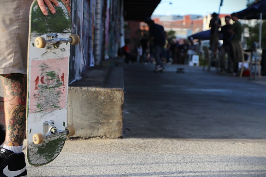 An unidentified man carries a skateboard next to his tattooed leg.