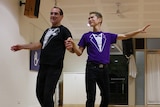John Cork and Steve Jeong dance the Cha Cha in a white-walled hall with a polished wood floor.