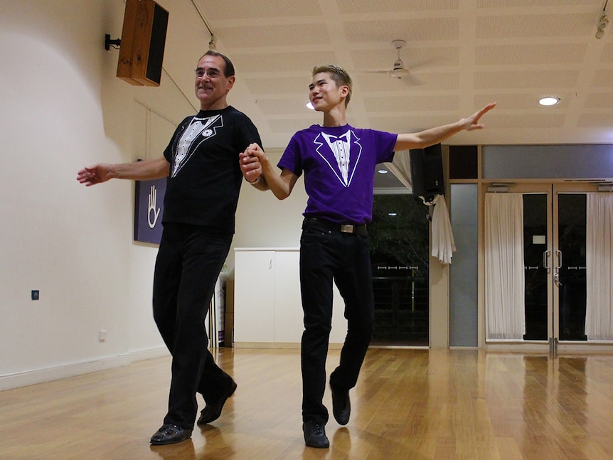 John Cork and Steve Jeong dance the Cha Cha in a white-walled hall with a polished wood floor.