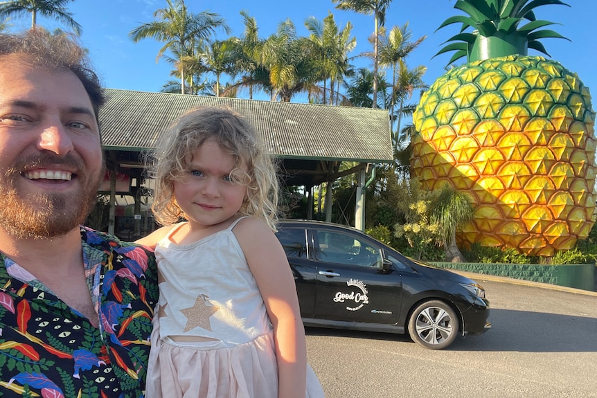 Blonde man and small girl with black car and giant pineapple in background.