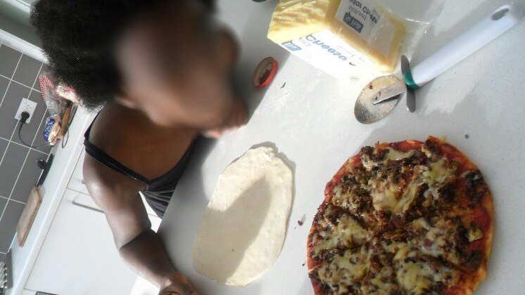 A girl leans over a table as she makes pizza, with a pizza base and a fully made pizza on the table.