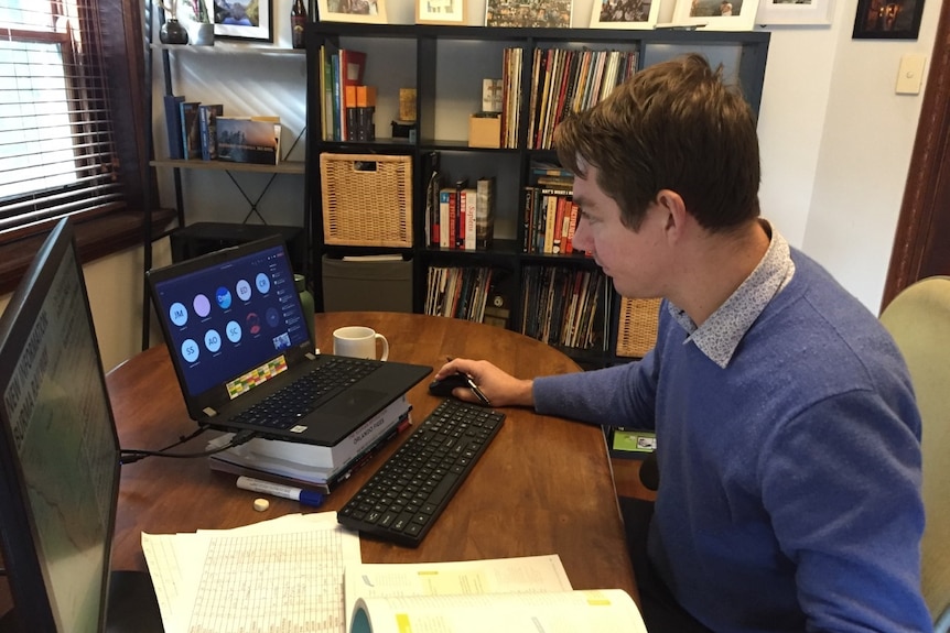 A man in a blue jumper sits at a table and works on a computer with history text books lying on the table.