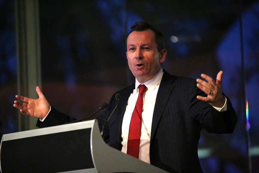 Mark McGowan standing at a podium wearing a suit, white shirt, red tie and speaking with his hands before a window at dusk
