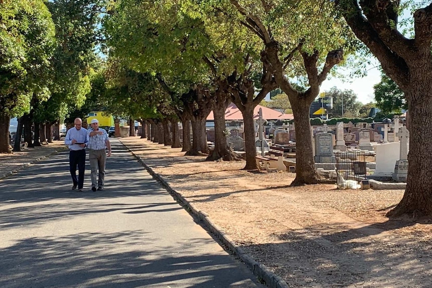 Two men walk down a road alongside a cemetery
