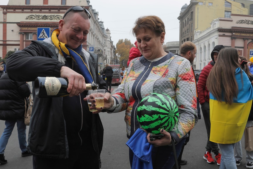 A man pours champagne into a cup for a woman as they stand on a street around people draped in the Ukrainian flag.