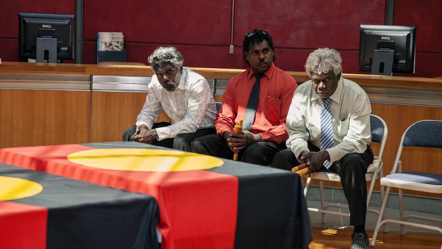 Three men are looking at boxes of bones covered with the indigenous flag.