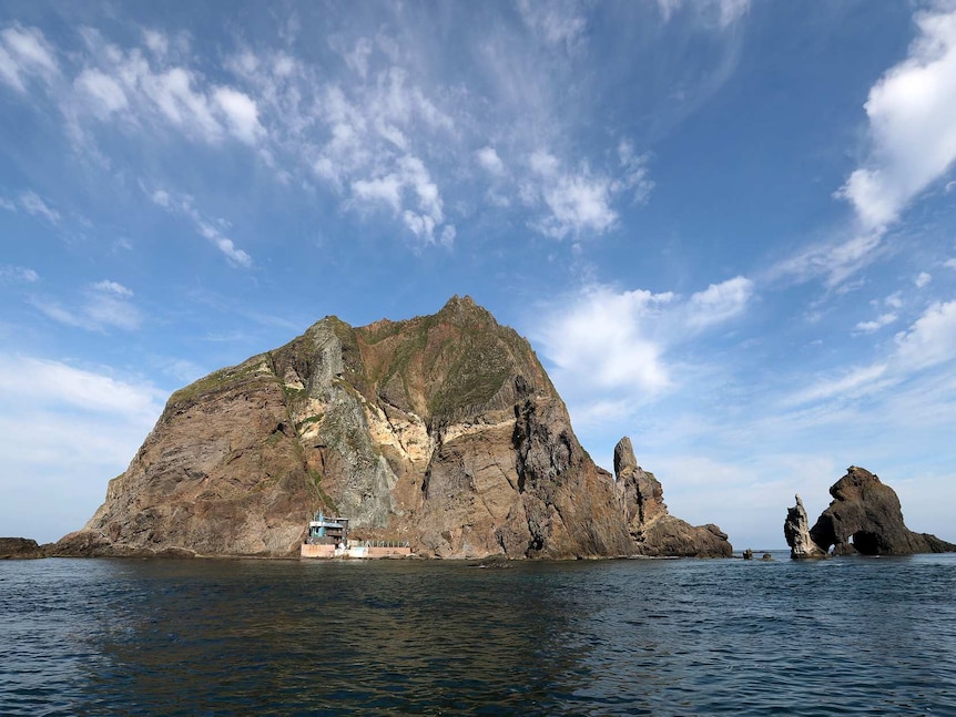 Looking out across a body of water, a rocky island is seen across the horizon line against a clear blue sky.