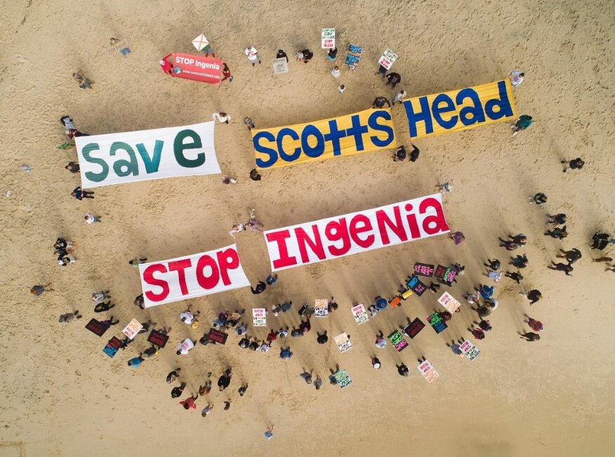 A drone shot of people gathering on a beach holding large signs that say 'Save Scotts head Stop Ingenia'