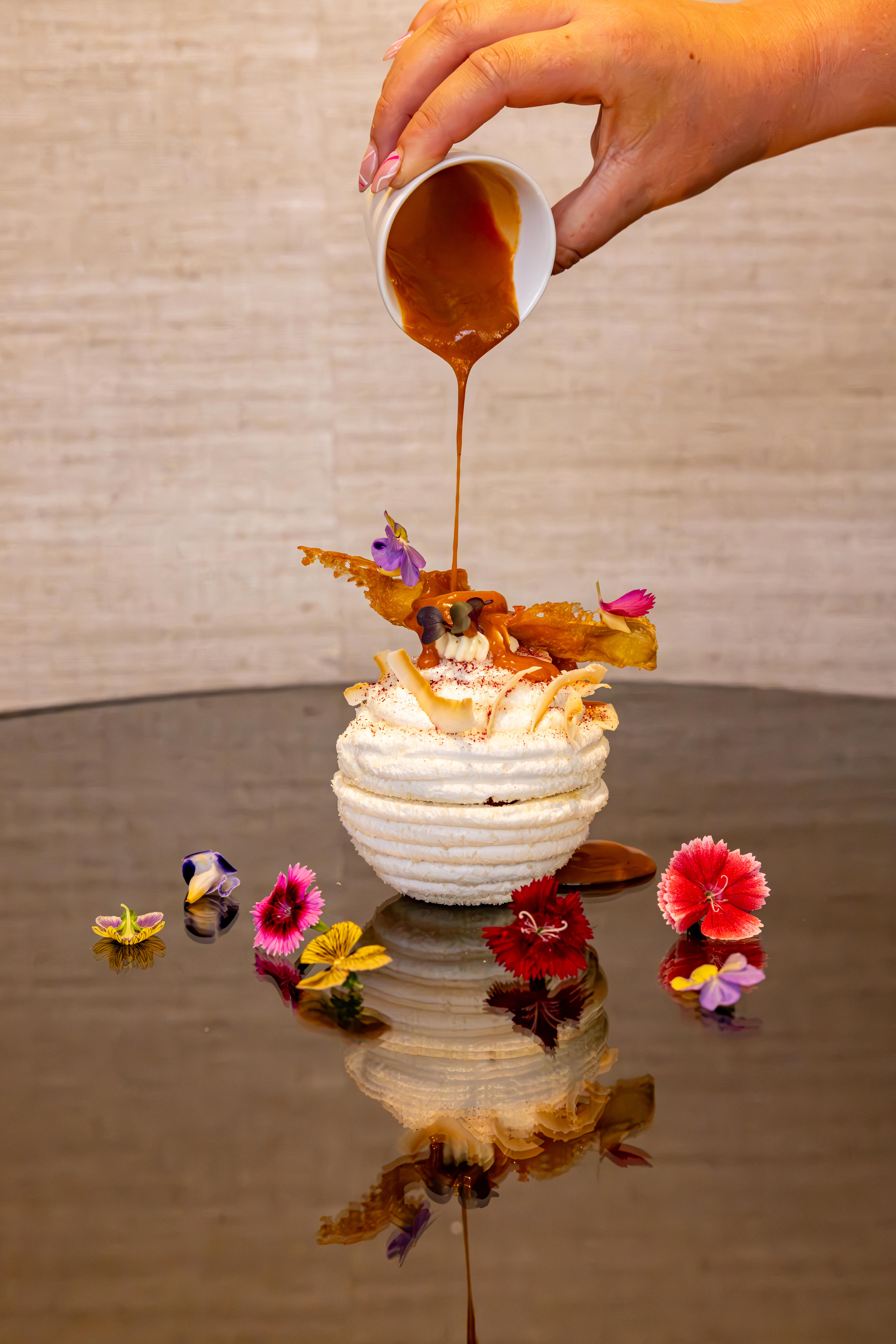 A hand pours syrup over a ball of white meringue topped with and surrounded by colourful flowers.
