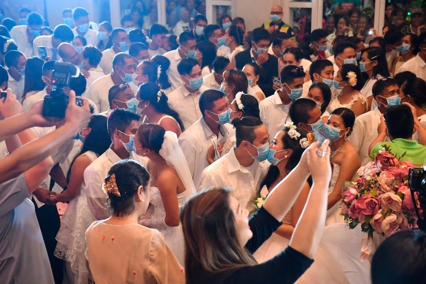 People take photos of couples in formal dress and wearing masks, who hug and kiss in a crowded hall.
