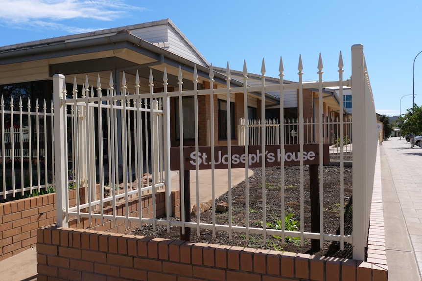 A brown house with a sign of St Joseph's House sits behind a beige fence with a clear blue sky.