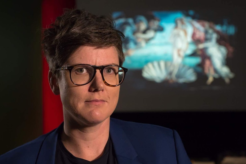 Hannah Gadsby standing in front of a screen featuring a projected image of Boticelli's painting 'The Birth of Venus'.