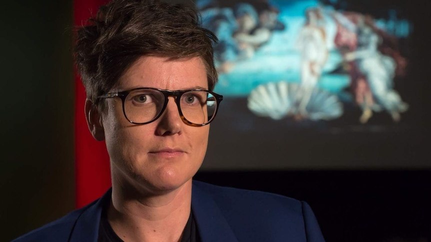 Hannah Gadsby standing in front of a screen featuring a projected image of Boticelli's painting 'The Birth of Venus'.