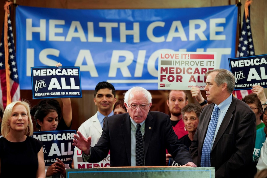 Bernie Sanders at a lectern surrounded by supporters holding "Medicare for all" posters