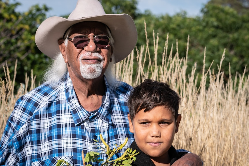 An older Indigenous Australian man wearing a cowboy hat with his hand on his grandson's shoulder.