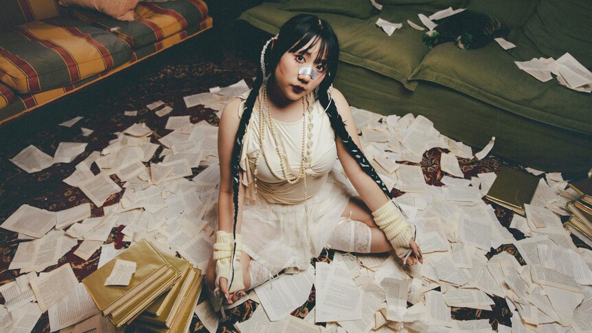 A woman sits on the floor in a white lace dress with book pages scattered around her