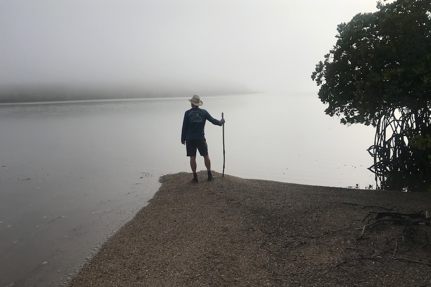 A man stands with a staff on the edge of a beach, looking out into the fog.