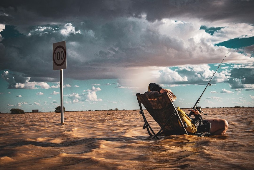 A man sits in a chair with his back to the camera, fishing rod in hand in muddy waters. There is a 100kph speed sign behind.