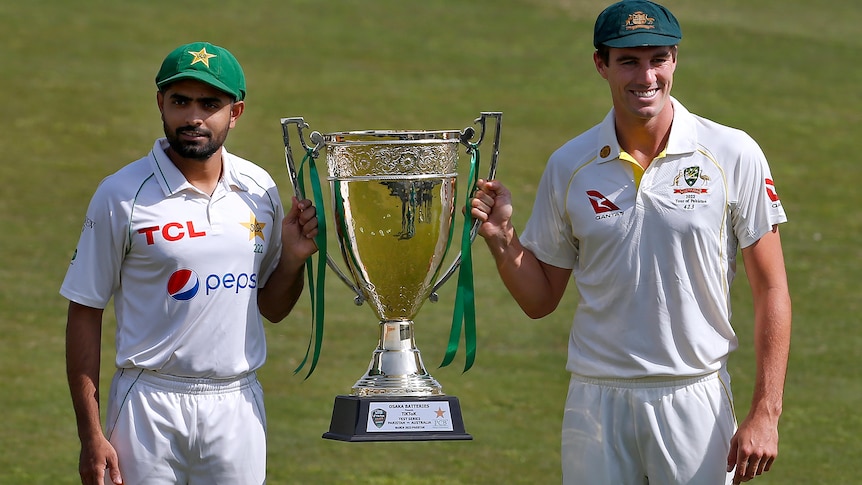 Babar Azam and Pat Cummins, wearing whites, smile while holding a large trophy together