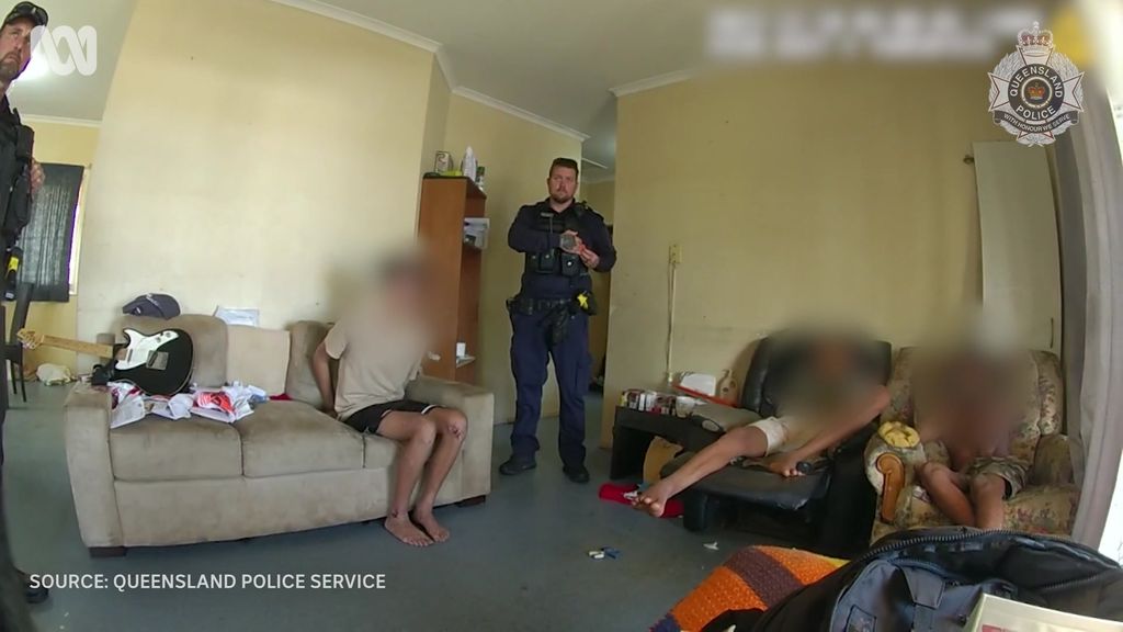 A police officer stands in a large living room. People whose faces are blurred sit on couches.