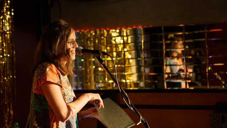 A young woman with glasses stands on a stage speaking into a mic and smiling, gold in the background